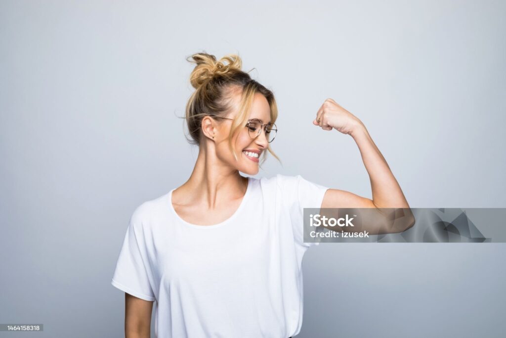 Happy beautiful woman flexing muscle while standing against gray background